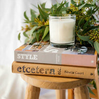 small CandleXchange candle sitting in front of eucalyptus branches and a pile of books on a stool